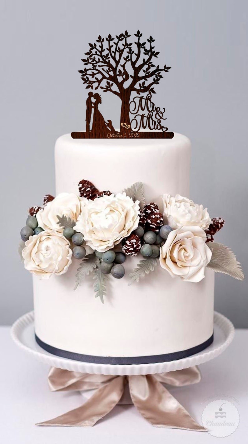 May include: A two-tiered white wedding cake with a wooden tree topper featuring a couple, a dog, and the text "Mr. & Mrs. October 1, 2022". The cake is decorated with white flowers, pine cones, and berries.