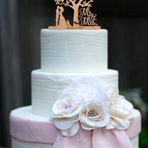 May include: A white wedding cake with a pink ribbon and flowers. The cake is topped with a wooden cake topper featuring a couple standing under a tree with the words "Mr. & Mrs." and the date "October 1, 2022".