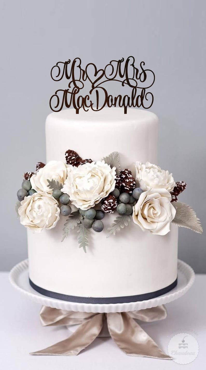 May include: A two-tiered white wedding cake decorated with white flowers, pine cones, and berries. The cake topper is a wooden sign that reads "Mr & Mrs MacDonald".