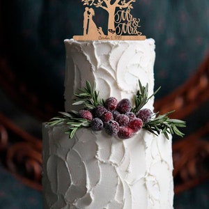 May include: A two-tiered white wedding cake with a wooden cake topper featuring a tree with two figures holding hands, a dog, and the text "Mr. & Mrs. October 1, 2022". The cake is decorated with sprigs of rosemary and red berries.