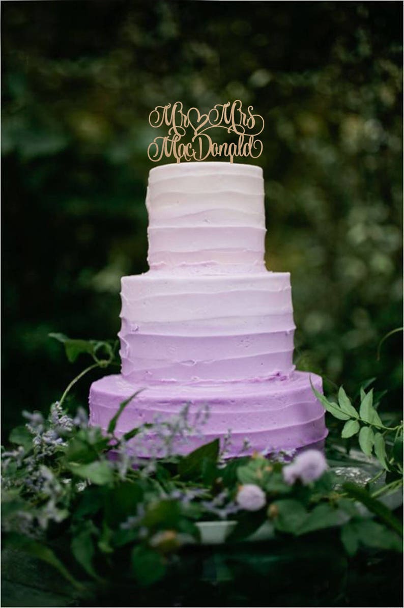 May include: A three-tiered ombre wedding cake with purple frosting. The cake is decorated with a wooden cake topper that says "Mr & Mrs Mac Donald".
