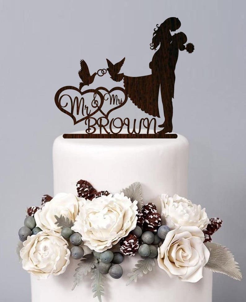 K&ouml;nnte beinhalten: Ein h&ouml;lzerner Hochzeitstortenaufsatz mit einer Silhouette eines sich umarmenden Paares, zwei V&ouml;geln mit einem Ring und dem Text "Mr. & Mrs. Brown".