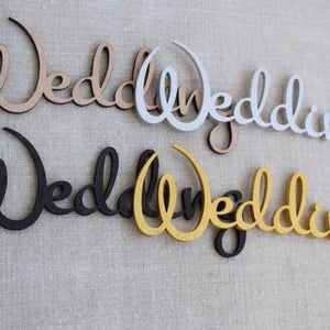 K&ouml;nnte beinhalten: Vier Holzschilder mit der Aufschrift "Wedding" in verschiedenen Farben: Naturholz, Wei&szlig;, Schwarz und Gold.
