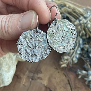 May include: A pair of silver-toned, textured earrings with a floral pattern. The earrings are held up by a silver hook. The earrings have a hammered appearance and are slightly irregular in shape. The background is a blurred wooden surface.