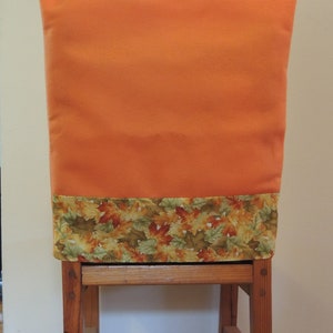 May include: An orange chair cover with a fall leaf pattern at the bottom. The cover is on a wooden chair with a simple design. The chair is made of wood and has a natural finish.