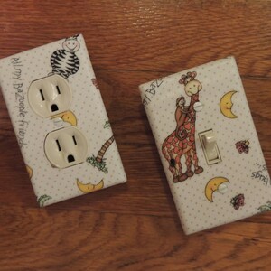 May include: Two white light switch covers with a decorative design. One cover features a giraffe and a monkey with the text "Sun, Fun, Friends". The other cover features a zebra and the text "All my Bazoople friends".