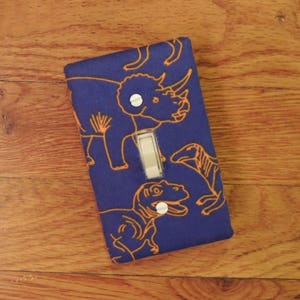 May include: A blue light switch cover with an orange dinosaur pattern. The dinosaurs include a triceratops, a stegosaurus, and a tyrannosaurus rex.