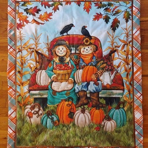 May include: A fall-themed decorative square featuring a red truck bed filled with pumpkins and scarecrows. The image includes pumpkins, corn stalks, and autumn leaves. The border has a plaid pattern in orange, white, and blue.