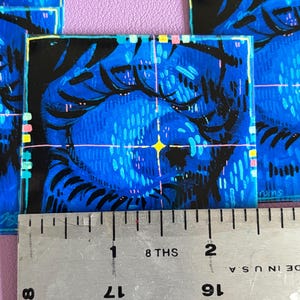 May include: Square art print featuring a blue and black illustration of a face with a yellow star. The artwork has a textured appearance with a ruler showing inches. The print is on a purple surface.