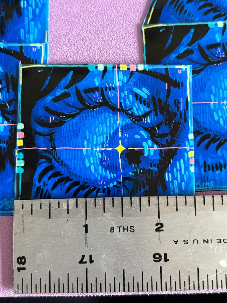 May include: Square art print featuring a blue and black illustration of a face with a yellow star. The artwork has a textured appearance with a ruler showing inches. The print is on a purple surface.