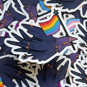 May include: A sticker featuring a dark blue crow with a rainbow pride flag. The crow is holding the flag with its beak and has a star on top. The sticker has a white background with glitter accents.