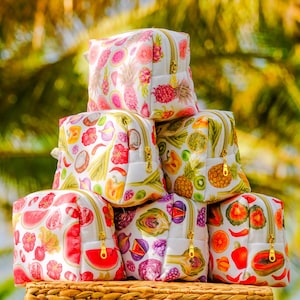 May include: A stack of six white cosmetic pouches featuring colorful fruit and vegetable patterns. Each rectangular bag has a gold-toned zipper closure. The prints include various produce like pineapples, watermelons, and peppers.