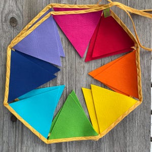 Puede incluir: Una guirnalda de banderines con banderas triangulares de fieltro en tonos violeta, azul oscuro, turquesa, verde, amarillo, naranja, rojo y magenta. Las banderas están unidas a una cinta de tela amarilla con un borde cosido en zigzag.