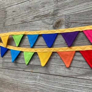 Może przedstawiać: Baner z filcu w kolorach tęczy z żółtym przeszyciem. Baner składa się z trójkątnych flag w kolorach: czerwony, pomarańczowy, żółty, zielony, niebieski, indygo i fioletowy.