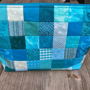 以下が含まれることがあります： さまざまな青とターコイズの色合いのパッチワークデザインのキルティングコスメティックバッグ。 バッグは長方形で、ジッパー開閉式で、星、水玉、幾何学模様など、さまざまなパターンを持つ正方形の生地で作られています。