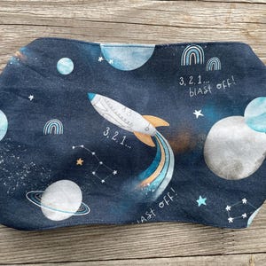 Könnte beinhalten: Ein marineblaues Stoffstück mit Weltraum-Motiv, einschließlich Rakete, Planeten, Sternen und Regenbogen. Der Text "3, 2, 1... blast off!" ist aufgedruckt. Das Objekt hat eine abgerundete Form.