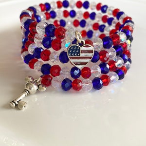 Pu&ograve; includere: Un braccialetto di perline rosso, bianco e blu con un ciondolo a forma di cuore che presenta la bandiera americana. Il braccialetto &egrave; avvolto attorno a una superficie bianca.
