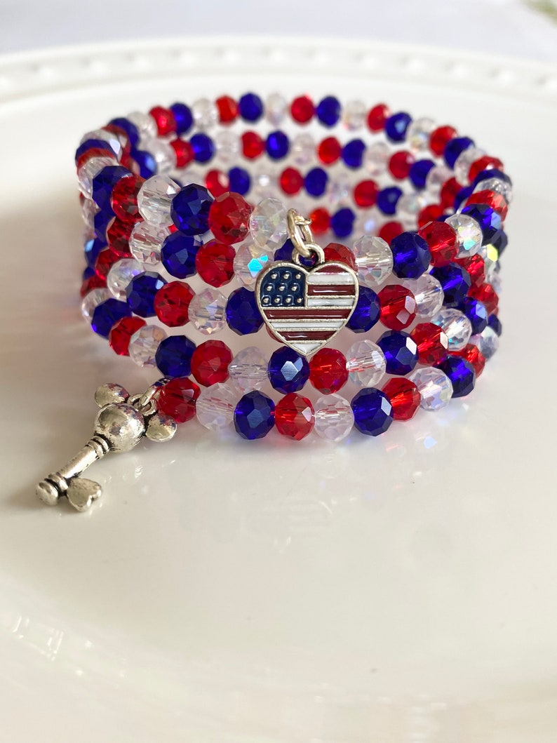 Pu&ograve; includere: Un braccialetto di perline rosso, bianco e blu con un ciondolo a forma di cuore che presenta la bandiera americana. Il braccialetto &egrave; avvolto attorno a una superficie bianca.