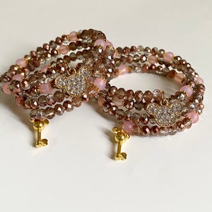 May include: Two gold key charm bracelets with pink and brown crystal beads. Each bracelet has a charm in the shape of a heart with rhinestones.