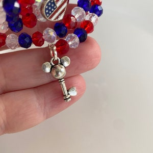 Pu&ograve; includere: Un braccialetto di perline rosso, bianco e blu con un ciondolo a forma di cuore argentato con una bandiera americana e un ciondolo a forma di chiave argentato con le orecchie di Topolino.