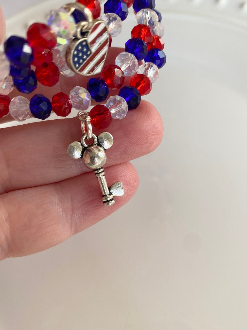 Pu&ograve; includere: Un braccialetto di perline rosso, bianco e blu con un ciondolo a forma di cuore argentato con una bandiera americana e un ciondolo a forma di chiave argentato con le orecchie di Topolino.