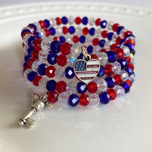 Pu&ograve; includere: Un braccialetto di perline rosso, bianco e blu con un ciondolo a forma di cuore argentato che presenta un design della bandiera americana. Il braccialetto &egrave; avvolto attorno a una superficie bianca.