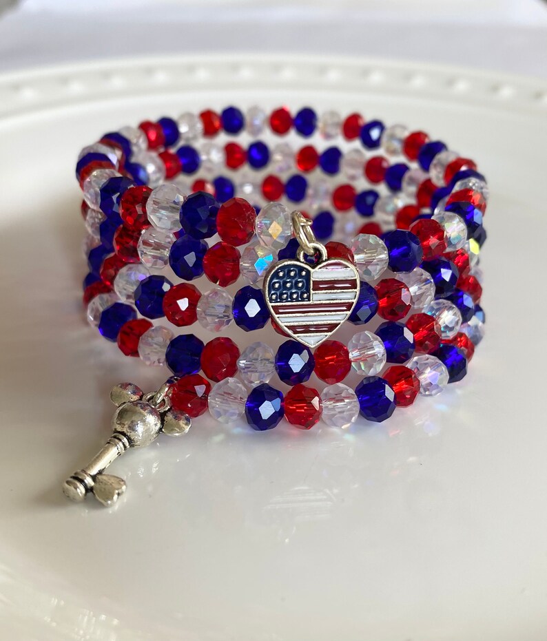 Pu&ograve; includere: Un braccialetto di perline rosso, bianco e blu con un ciondolo a forma di cuore argentato che presenta un design della bandiera americana. Il braccialetto &egrave; avvolto attorno a una superficie bianca.
