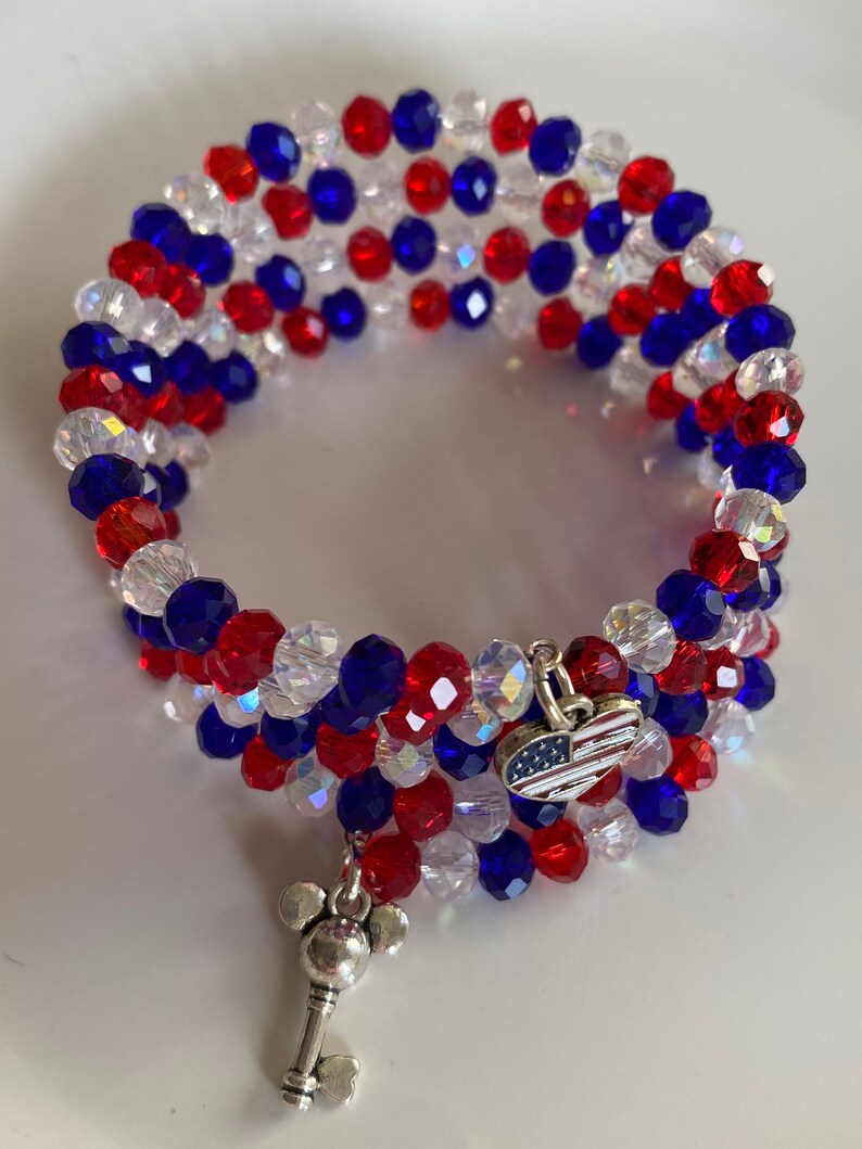 Pu&ograve; includere: Un braccialetto di perline rosso, bianco e blu con un ciondolo d'argento a forma di chiave con le orecchie di Topolino e un ciondolo d'argento a forma di cuore con un design di bandiera americana.
