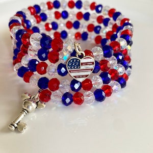Pu&ograve; includere: Un braccialetto di perline rosso, bianco e blu con un ciondolo d'argento a forma di cuore con un design di bandiera americana. Il braccialetto &egrave; avvolto attorno a una superficie bianca.