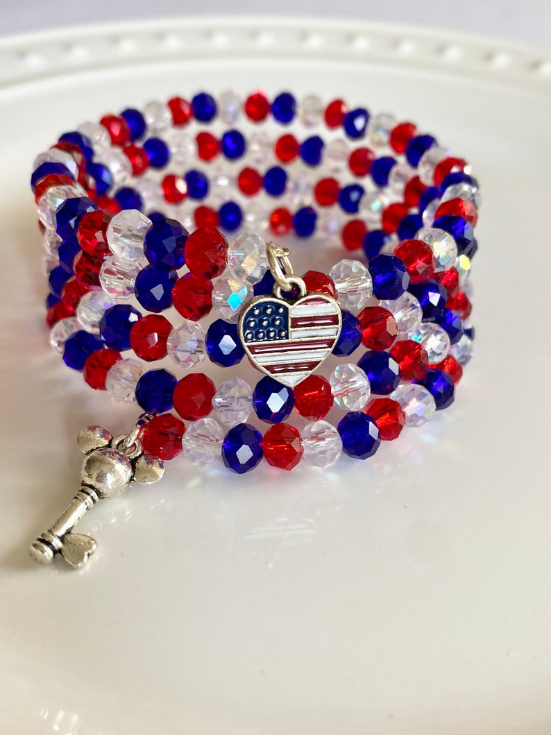 Pu&ograve; includere: Un braccialetto di perline rosso, bianco e blu con un ciondolo d'argento a forma di cuore con un design di bandiera americana. Il braccialetto &egrave; avvolto attorno a una superficie bianca.