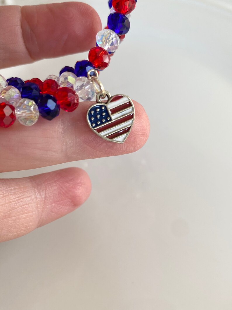 Pu&ograve; includere: Un braccialetto di perline rosso, bianco e blu con un ciondolo a forma di cuore argentato con un design di bandiera americana.