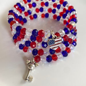 Pu&ograve; includere: Un braccialetto di perline rosso, bianco e blu con un ciondolo a forma di cuore con la bandiera americana e un ciondolo a forma di chiave.