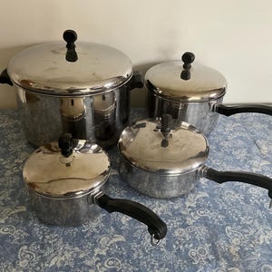 May include: A set of four stainless steel cooking pots with matching lids. Each pot features a polished silver finish, black heat-resistant handles, and black knobs on the lids. The handles include small metal loops for hanging storage.