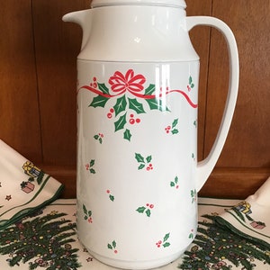 May include: White insulated carafe with a lid and handle, decorated with a red ribbon, green holly leaves, and red berries. The carafe is part of a Christmas-themed set, with matching tablecloths visible in the background.