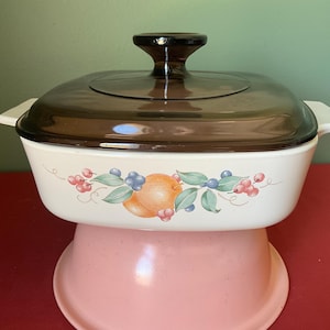 May include: A white casserole dish with a brown glass lid and a pink base. The dish features a floral design with fruit and berries.