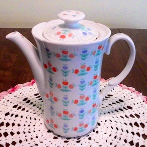 Puede incluir: Cafetera de cerámica blanca con un estampado floral en rojo, azul y verde. La cafetera tiene un asa curva, un pico y una tapa con un diseño floral a juego. Se encuentra sobre un tapete de ganchillo blanco con borde rosa.