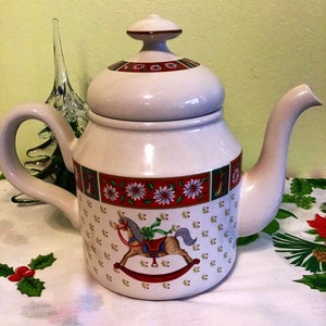 Op de afbeelding: Witte keramische theepot met een rood, groen en wit kerstthema. De theepot heeft een schommelpaard met een rood zadel en een groene strik. De deksel heeft een rood en wit bloemenpatroon.