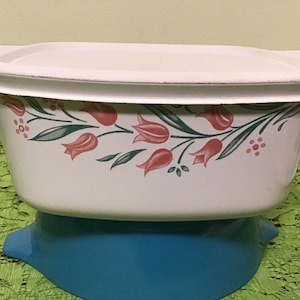 May include: White ceramic baking dish with a floral design featuring pink tulips and green leaves. The dish has a matching lid and sits on a blue base.