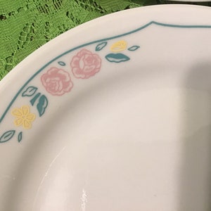Könnte beinhalten: Weißer Teller mit floralem Design. Der Rand hat eine türkisfarbene Kontur mit rosa Rosen, gelben Blumen und türkisfarbenen Blättern. Der Teller besteht aus einem haltbaren Material.