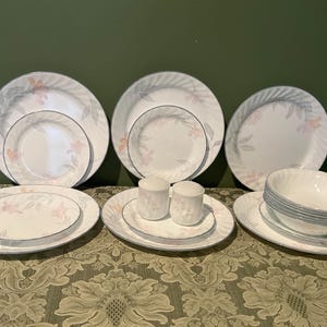 May include: A complete dinnerware set featuring white plates, bowls, and saucers with a floral design in pink and gray. The set includes dinner plates, salad plates, bowls, and salt and pepper shakers. The rims have a blue trim.