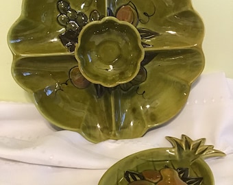 Puede incluir: Un juego de servir de cerámica verde. La gran fuente en forma de hoja tiene cuatro secciones y un cuenco central, decorado con uvas y frutas pintadas. También se incluye un plato más pequeño en forma de piña con diseños de frutas.