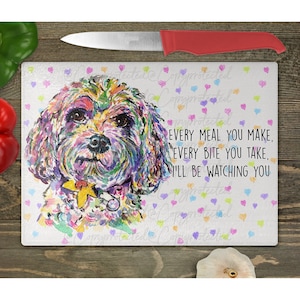 May include: A white cutting board with a colorful painting of a dog's face. The background is covered in small pink, purple, blue, and green hearts. The text "EVERY MEAL YOU MAKE, EVERY BITE YOU TAKE, I'LL BE WATCHING YOU" is printed on the board.