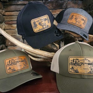 May include: Four trucker hats with a landscape design. The hats are black, gray, and olive green with a mesh back and a patch featuring a mountain and tree scene. The patches are rectangular and have a brown leather-like appearance.