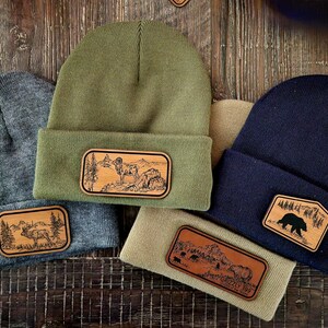 May include: Four knit beanies with different colored fabric and leather patches featuring various wildlife designs. The patches are rectangular and have a brown woodgrain texture. The beanies are arranged in a semi-circle on a wooden surface.