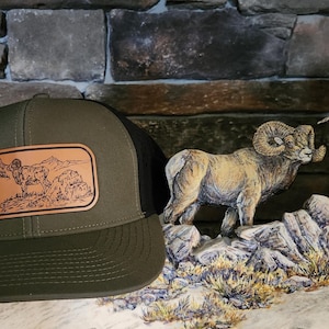 May include: A green and black trucker hat with a brown leather patch featuring a mountain scene with a bighorn sheep. The hat is sitting on a wooden surface with a carved bighorn sheep figurine.