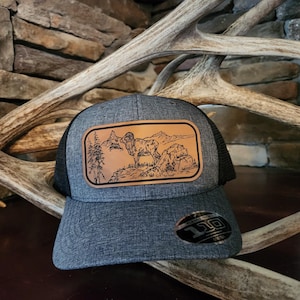 May include: A gray and black trucker hat with a brown leather patch featuring a detailed illustration of a bighorn sheep in a mountain landscape.