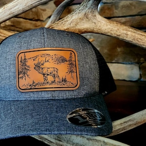 May include: A gray and black trucker hat with a brown leather patch featuring a detailed illustration of an elk in a mountain landscape. The patch is stitched onto the front of the hat.
