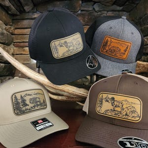 May include: Four baseball caps with mesh backs and rectangular patches featuring outdoor scenes. The caps are black, gray, tan, and brown. The patches depict mountains, trees, and wildlife in a rustic style.