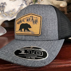 May include: A gray and black mesh trucker hat with a wood patch featuring a black bear silhouette in a forest scene. The patch has the text "Flexfit Tech 110 One Ten".