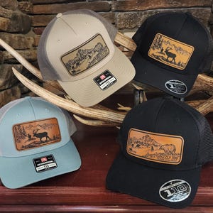 May include: Four trucker hats with mesh backs and leather patches. The hats are in tan, black, and blue-green. The patches feature mountain and wildlife scenes. One patch has the text "Grizzly 399".